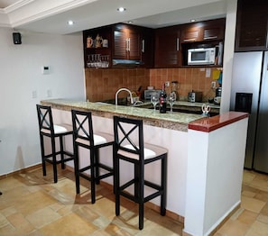 Uma cozinha com bancada de granito, quatro banquetas, geladeira de aço inoxidável e armários de madeira.