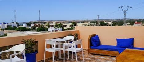 A rooftop terrace with a table and chairs, a wooden bench with blue cushions, and a view of the cityscape.