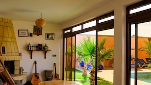 A room with a fireplace, a dining table, and a view of a pool and garden. - Dar Iziki (Sidi Kaouki)