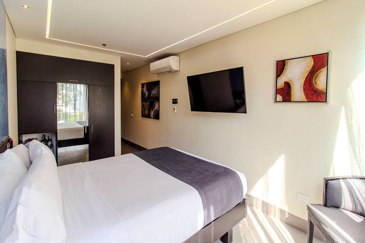 Una habitación de hotel moderna con una cama grande, un televisor de pantalla plana montado en la pared y un ventanal que proporciona luz natural.