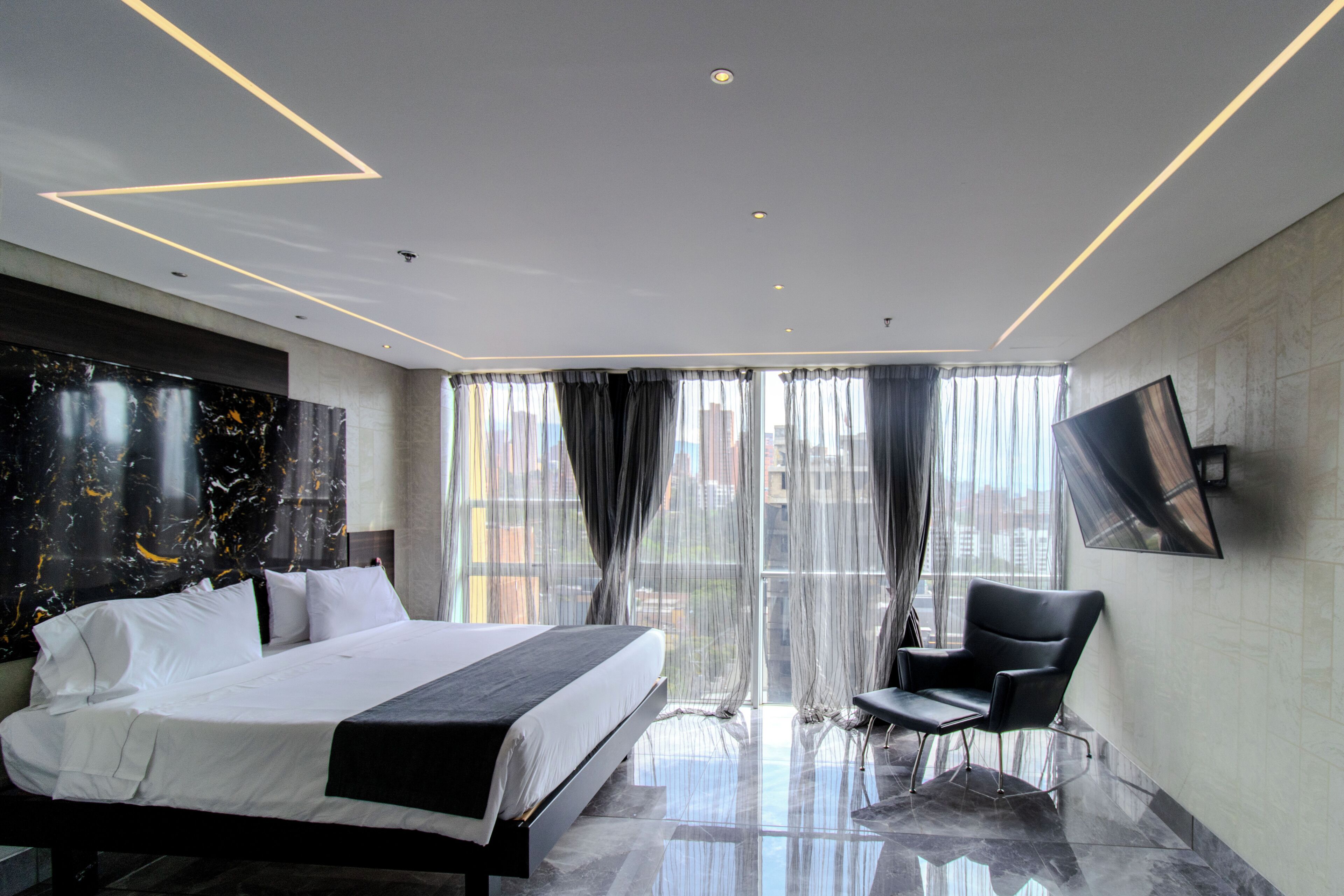 A modern hotel room with a large bed, a chair, a TV, and a city view.