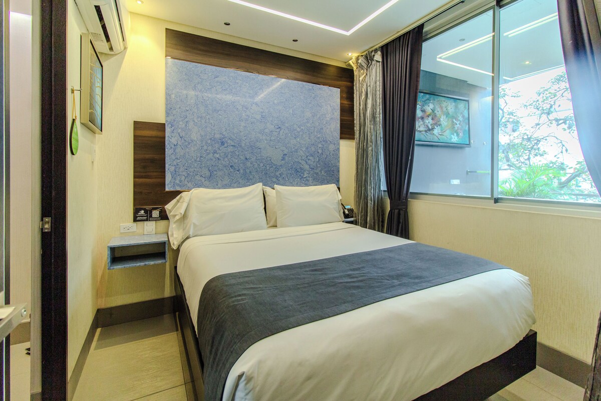 A hotel room with a large bed, a nightstand, a wall-mounted air conditioner, a window with curtains, and a painting.