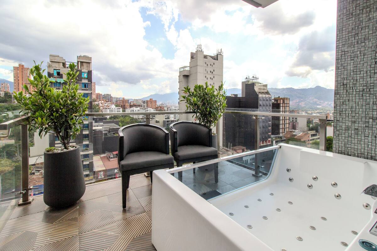 Un balcón con bañera de hidromasaje cerrada con vidrio, dos sillas y plantas en macetas.