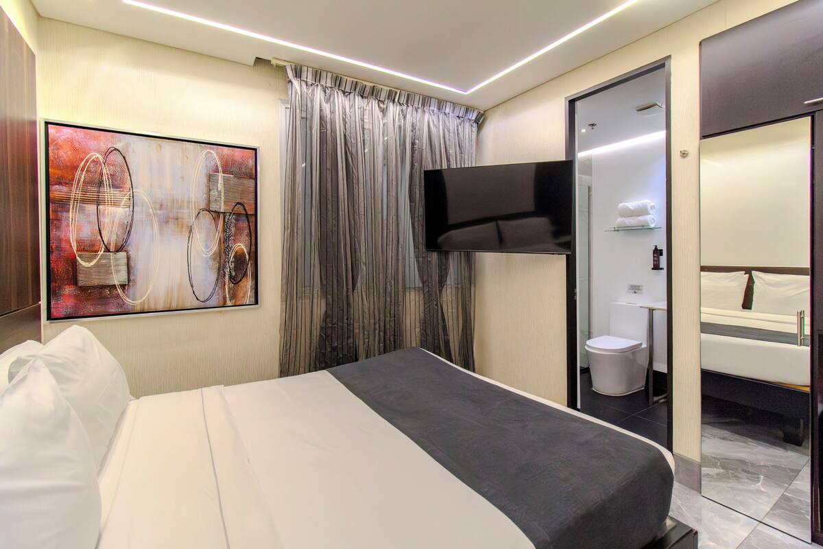 Habitación de hotel moderna con cama, televisor de pantalla plana, un cuadro abstracto grande, baño con espejo y retrete, y cortina.