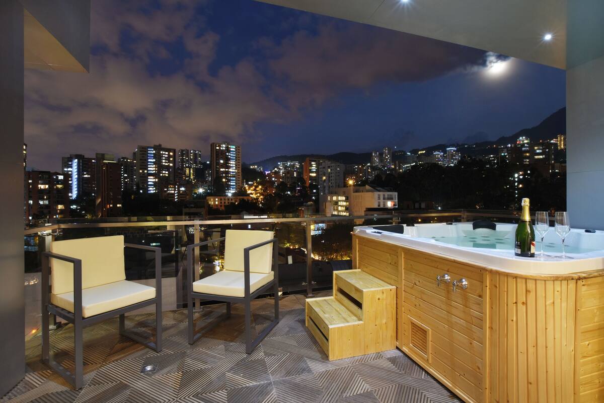 Terraza en la azotea con jacuzzi, dos sillas y vistas a la ciudad al atardecer.
