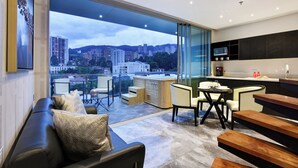 A modern living room with a large window, a sofa, a dining table, and a kitchen area with a microwave. - Haven, A Design Hotel (Medellín)