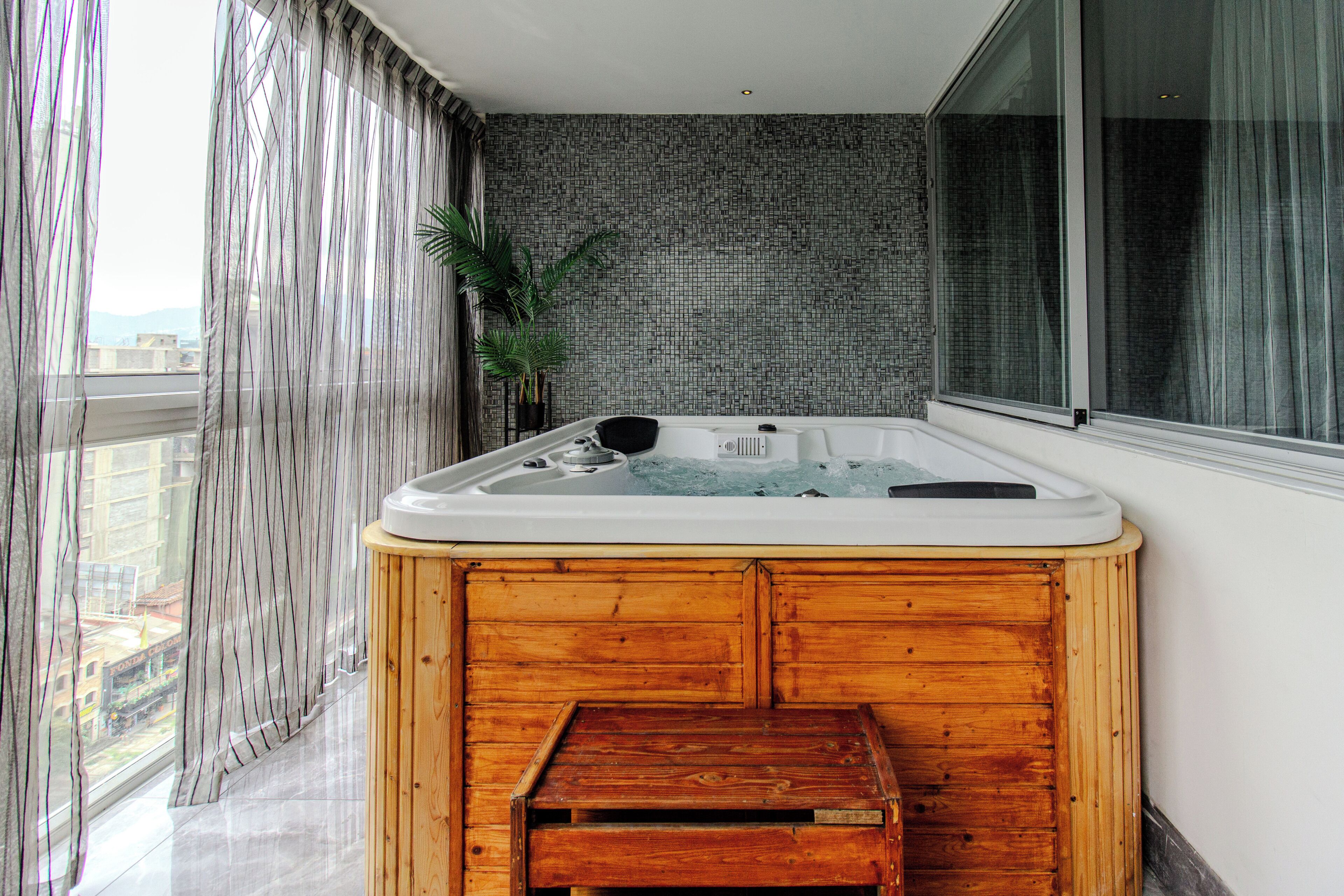 Un jacuzzi en una esquina con un banco de madera, una planta en maceta y vistas a edificios a través de amplios ventanales.