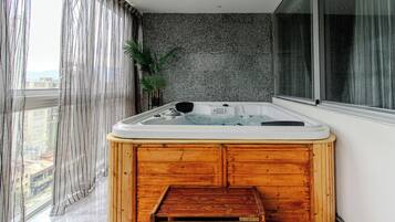 Un jacuzzi en una esquina con un banco de madera, una planta en maceta y vistas a edificios a través de amplios ventanales.