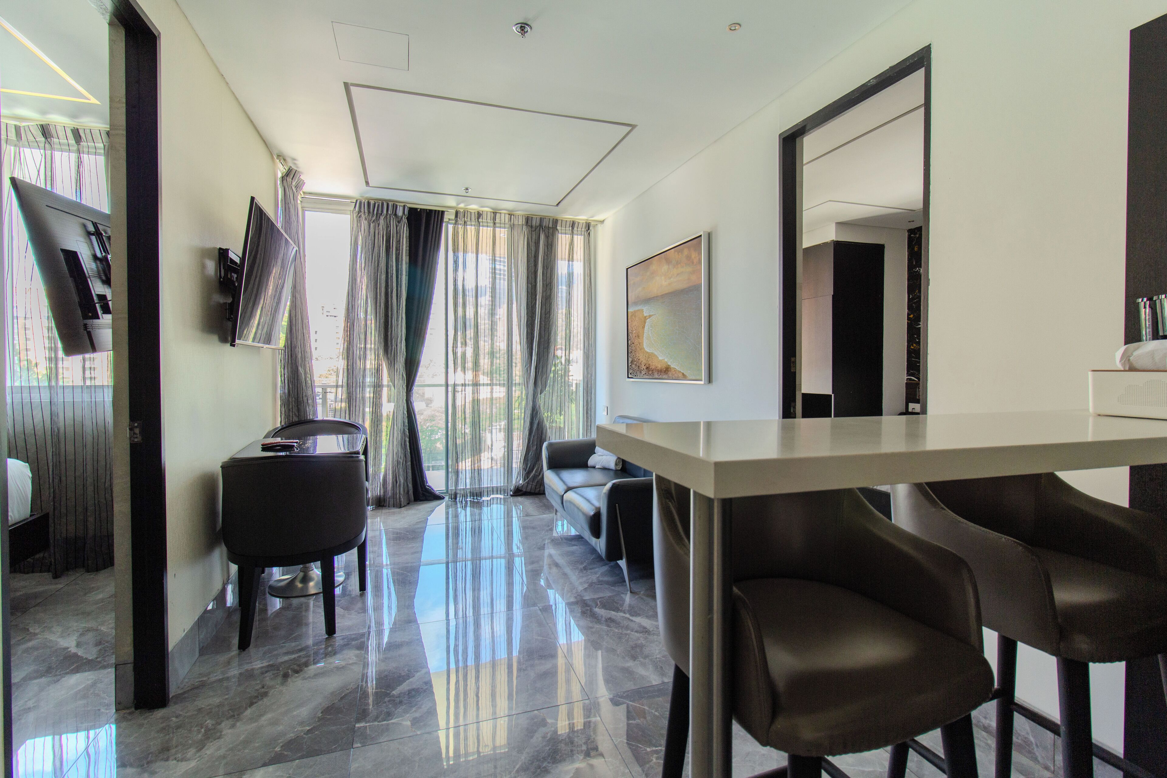 Une chambre d’hôtel moderne dotée d’une grande fenêtre, d’un téléviseur à écran plat fixé au mur, d’un canapé, d’une table à manger avec des chaises et d’un grand miroir.