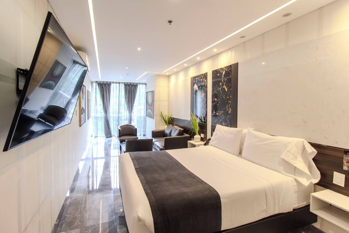Una habitación de hotel moderna con una cama grande, un televisor montado en la pared, una zona de estar con sillas y un ventanal con cortinas.