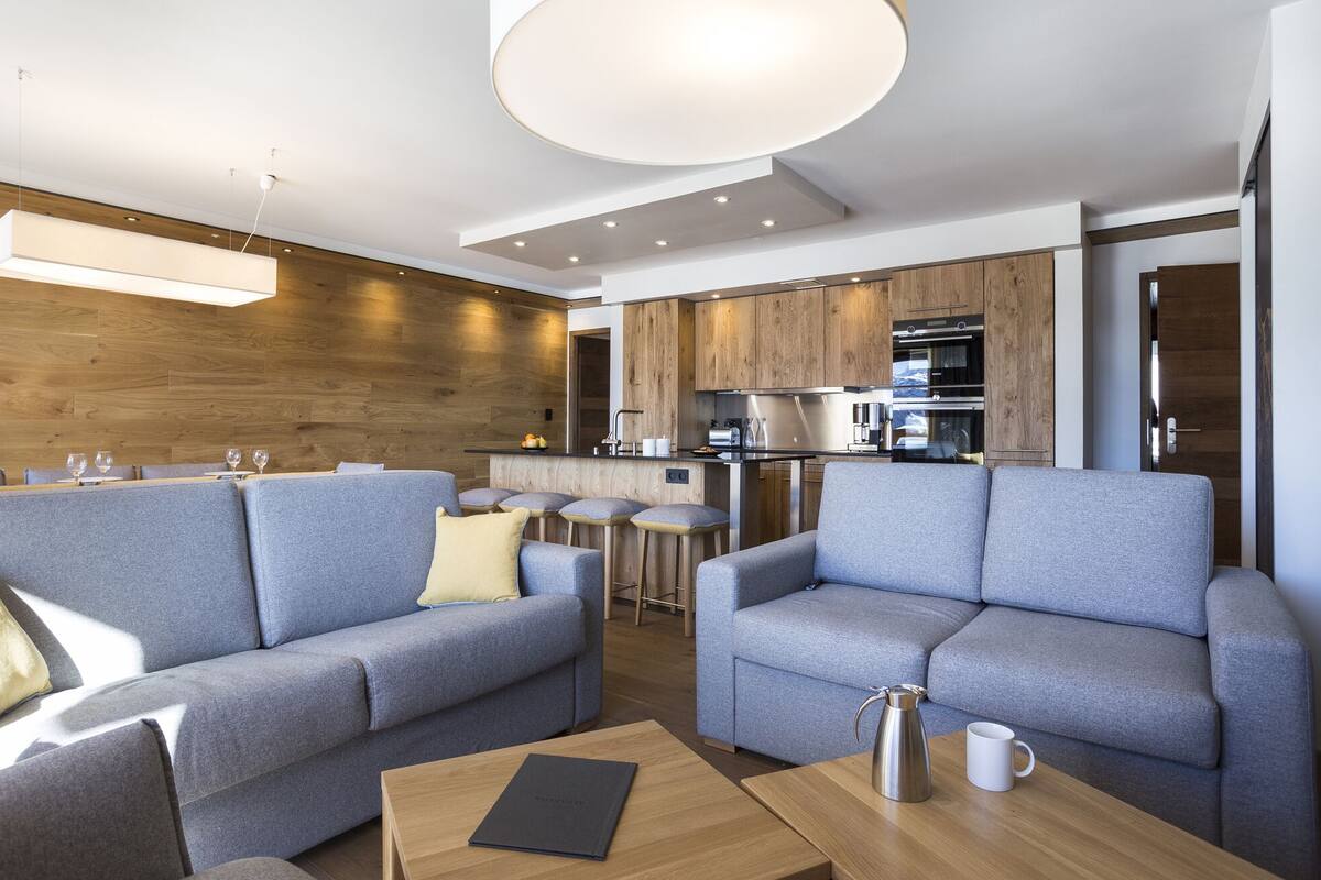 A modern living room with grey sofas, a coffee table, and a kitchen area with wooden cabinets and stainless steel appliances.