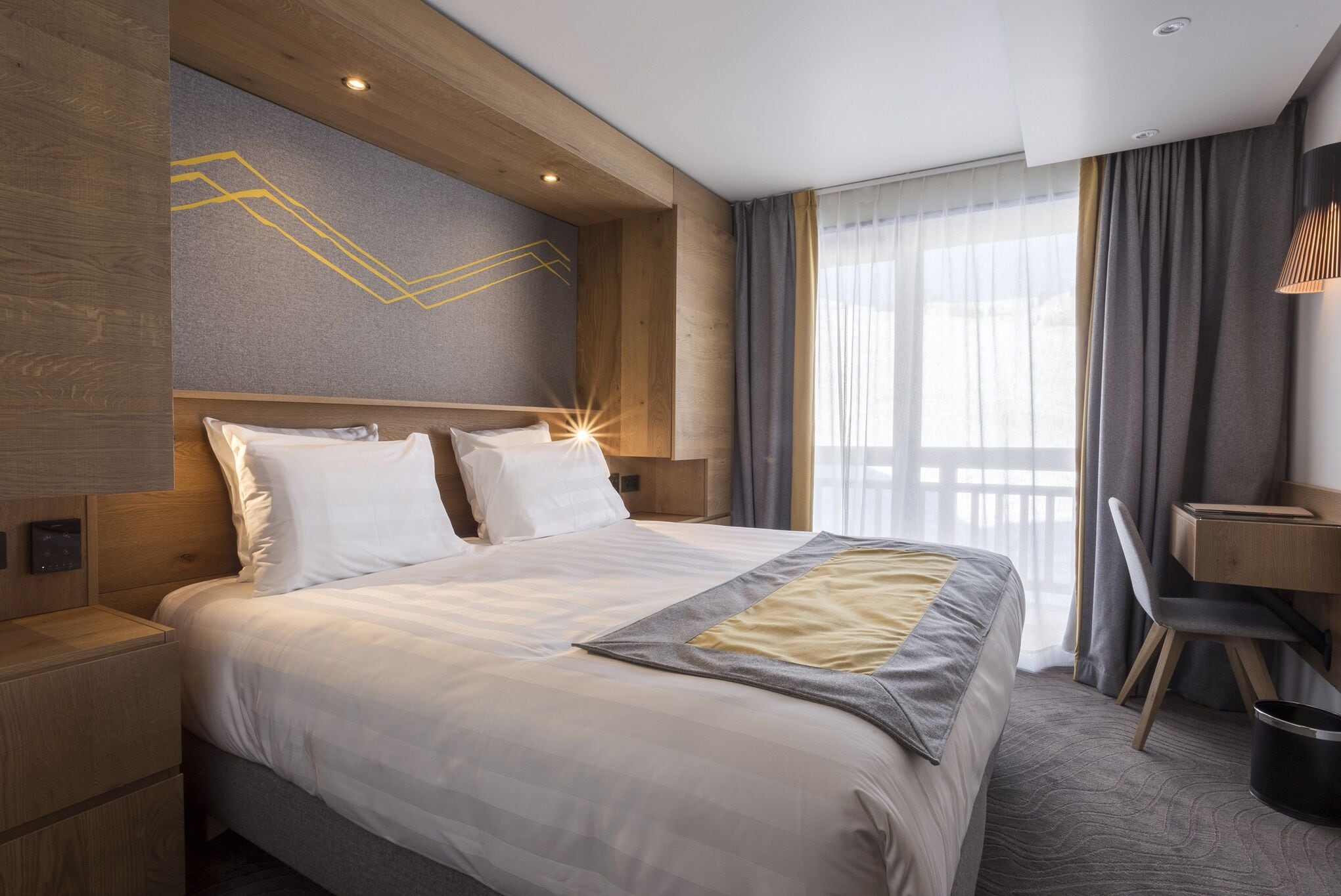 A modern hotel room with a large bed, wooden headboard, a desk, and a view of the outdoors.