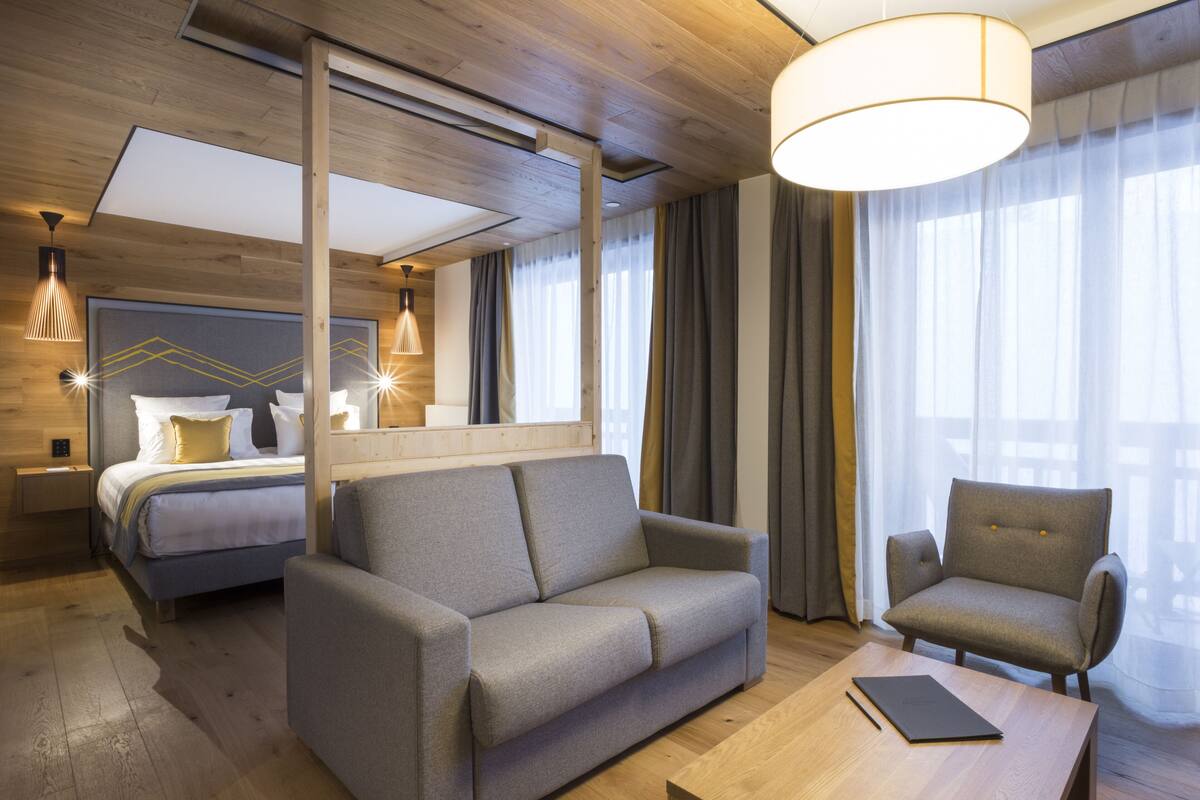 A modern hotel room with a bed, sofa, armchair, and a wooden coffee table.