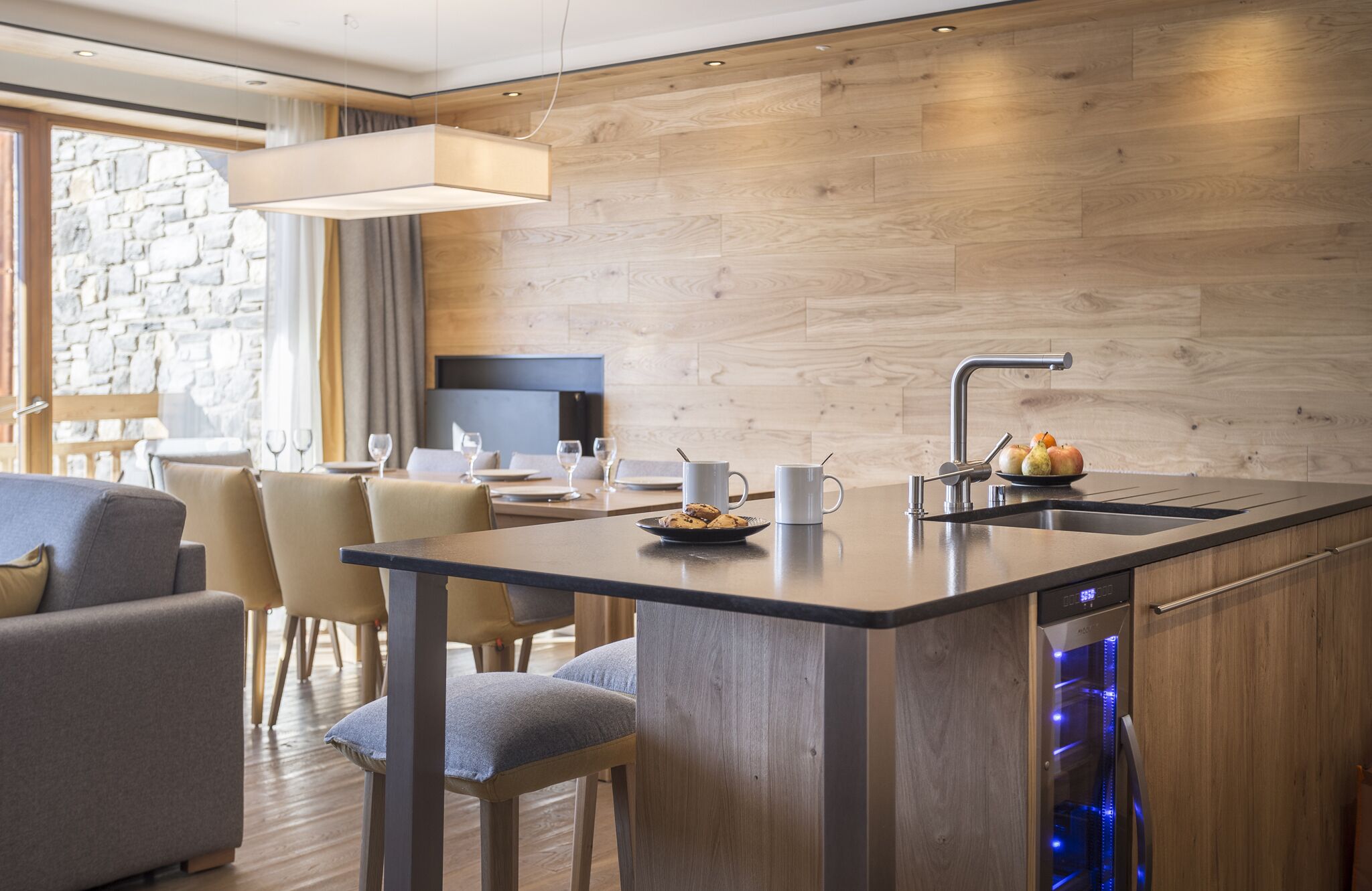 A modern kitchen with a central island, built-in dishwasher, and a stone wall in the background.