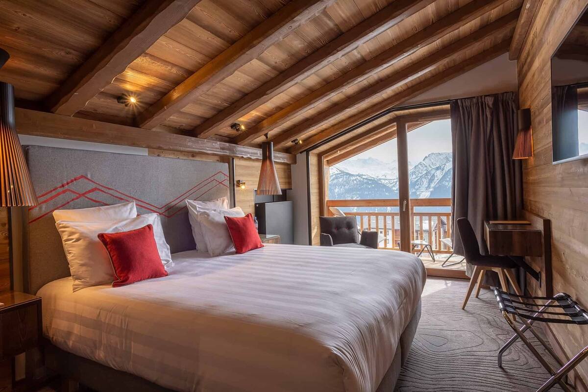 A hotel room with a large bed, a desk, and a view of mountains through a large window.