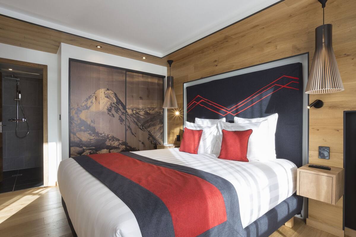 A modern hotel room with a large bed, wooden paneling, a mountain artwork on the wall, and a nightstand with a remote control.