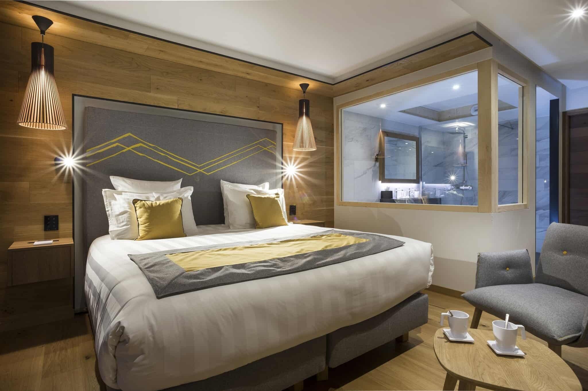 A modern hotel room with a large bed, bedside tables, a seating area with two chairs, and a large window offering a view of the city.