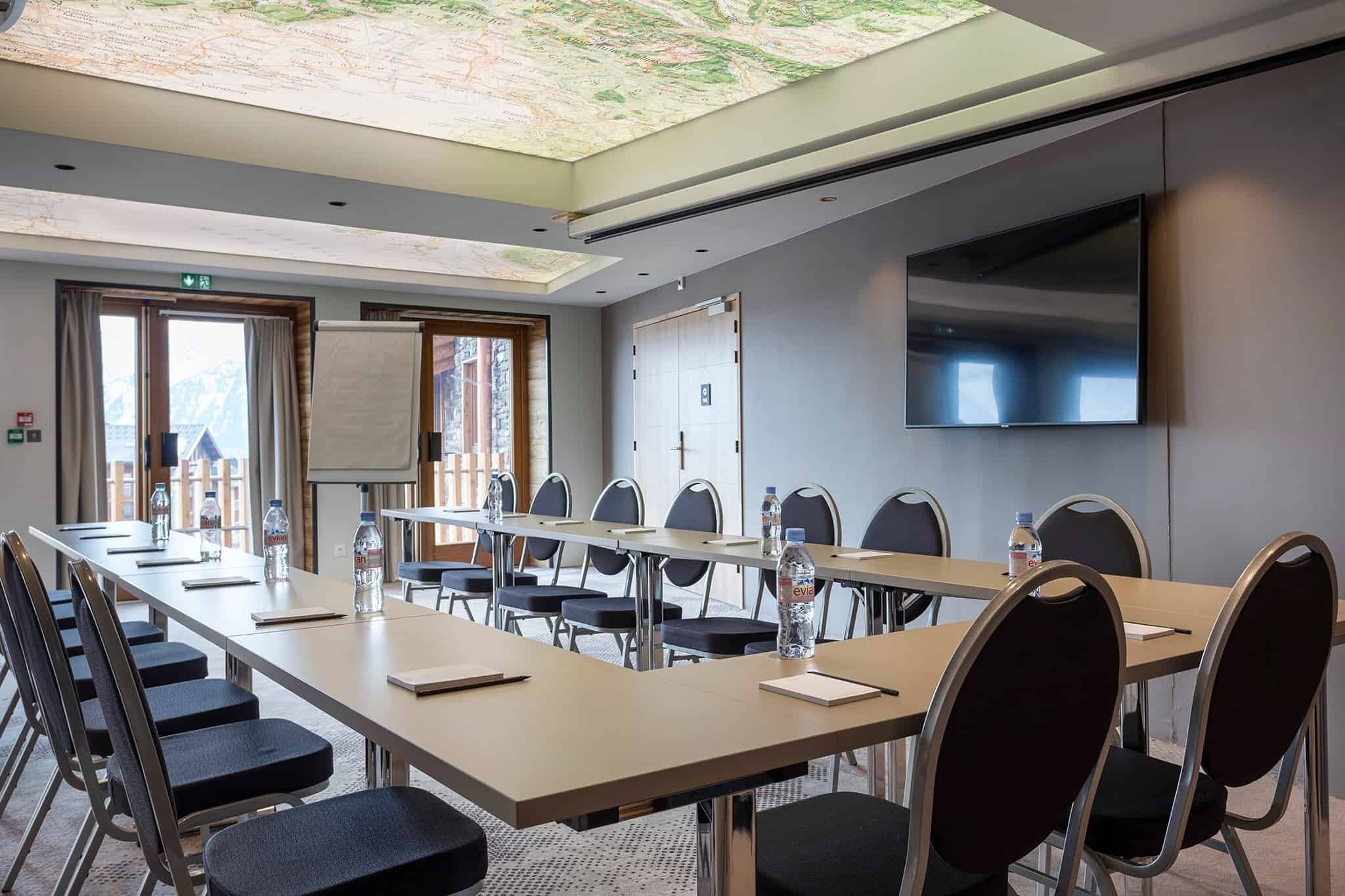 Un salón de conferencias con una larga mesa, sillas, una pantalla de proyector y un mapa en el techo.
