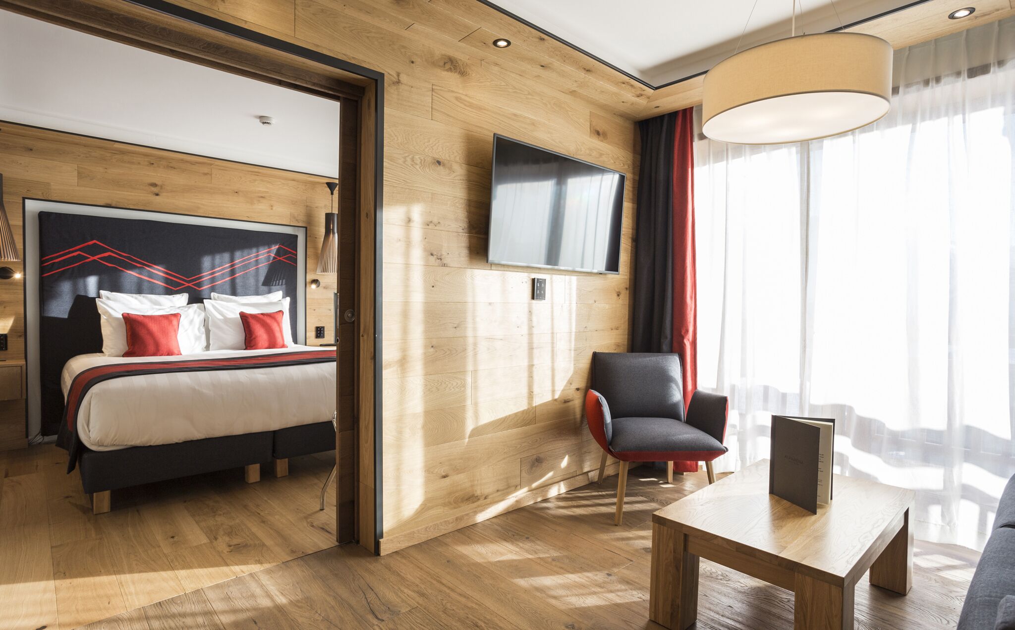 Een moderne hotelkamer met houten vloer, een bed met rode en witte beddengoed, een zwarte stoel, een houten tafel, een aan de muur bevestigde televisie en een groot raam met gordijnen.