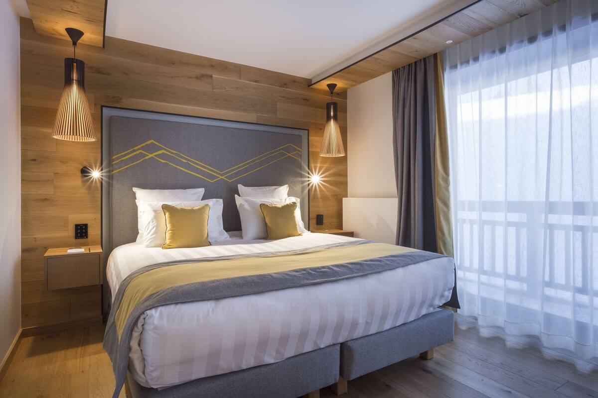 A modern hotel room with a large bed, wooden headboard, two bedside tables, and a large window with curtains.