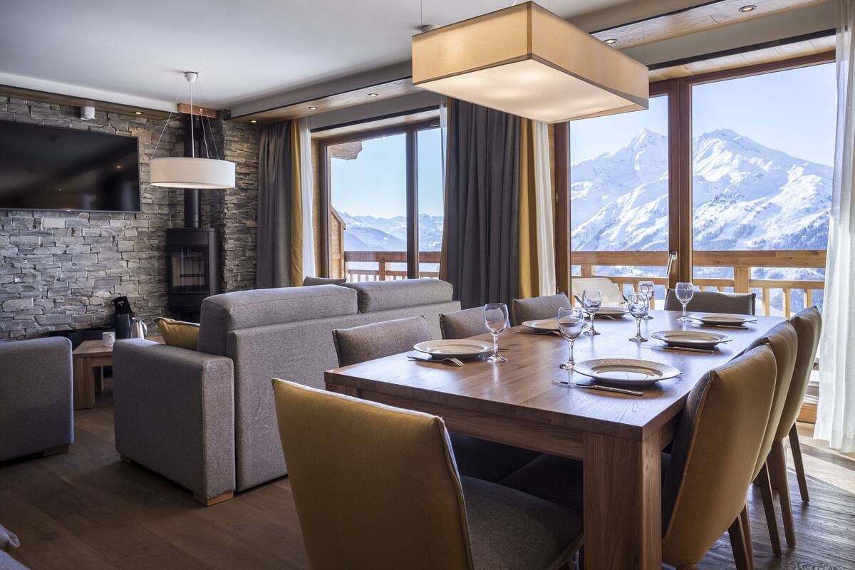 A modern dining area with a wooden table set for four, a stone fireplace, and a large window offering a mountain view.