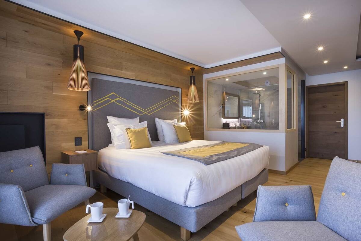 A modern hotel room with a large bed, two grey armchairs, a small table with a cup, and a large mirror on the wall.