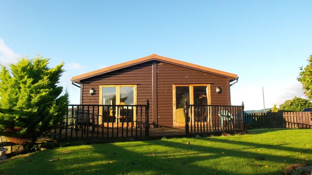 A small, single-story wooden cabin with a fenced yard, large windows, and a door. - Greenacres Holiday Cabins (St. Andrews)