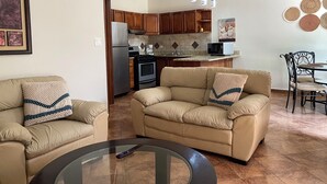 Un salón con sofás color beige, una mesa de centro de cristal y una cocina al fondo.