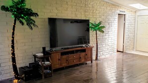 Sala de estar con un televisor de pantalla plana grande, mueble de entretenimiento de madera y dos palmeras artificiales con luces verdes.