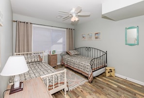 A bedroom with two beds, a ceiling fan, a window with blinds, a bedside table, a lamp, a mirror, and a rug.