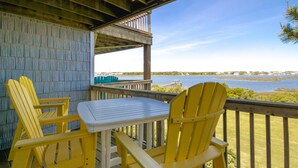 A wooden deck with a table and two chairs, overlooking a body of water with a distant shoreline. - Island In The Sun - 3BR Townhouse with Ocean & Sound Views! (North Topsail Beach)