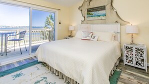 A bedroom with a large bed, a view of the sea through a sliding glass door, a decorative headboard, and a bedside table with a lamp. - Island In The Sun - 3BR Townhouse with Ocean & Sound Views! (North Topsail Beach)
