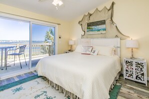 A bedroom with a large bed, a view of the sea through a sliding glass door, a decorative headboard, and a bedside table with a lamp. - Island In The Sun - 3BR Townhouse with Ocean & Sound Views! (North Topsail Beach)