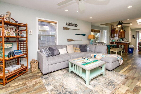 A living room with a sectional sofa, a coffee table, a bookshelf, and a wall-mounted sign reading 'SEASIDE'.