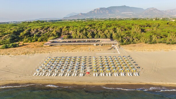 Une vue aérienne d’une plage avec des rangées de chaises longues et de parasols, un bâtiment doté d’un toit, et un paysage montagneux en arrière-plan.