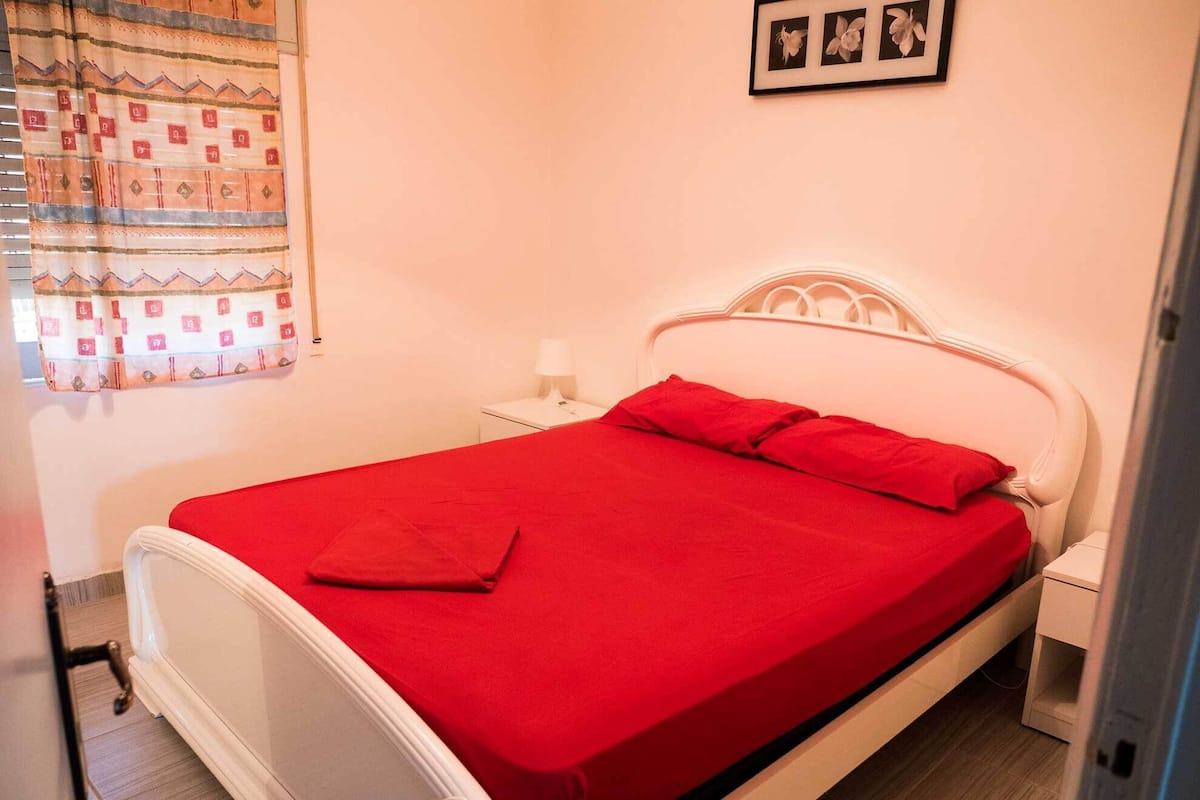 Ein Zimmer mit einem Einzelbett, einem weißen Kopfteil, einer roten Bettdecke und einem kleinen Nachttisch mit Lampe.