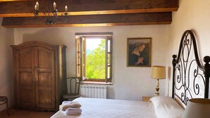 Een slaapkamer met een houten garderobe, een bed met witte lakens, een raam met uitzicht op groen, een schilderij aan de muur en een kroonluchter.
