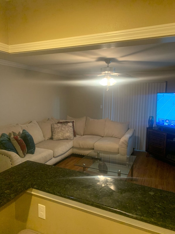 A living room with a sectional sofa, a glass coffee table, a ceiling fan, and a television displaying a sports game.
