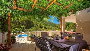 Un espace extérieur abrité, aménagé pour les repas, avec une table pour deux, une piscine en arrière-plan et une végétation luxuriante qui entoure l’endroit.