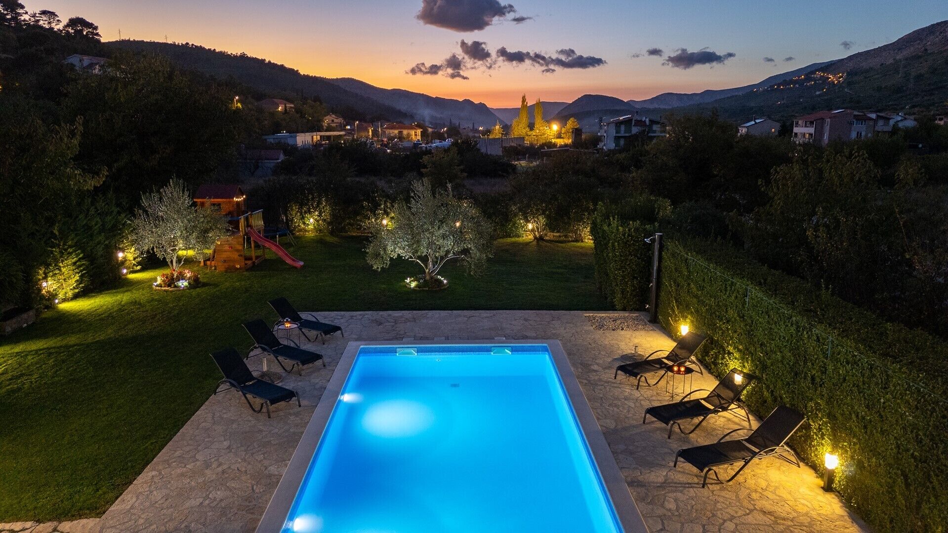 A rectangular swimming pool with a stone deck, surrounded by lounge chairs and small trees.