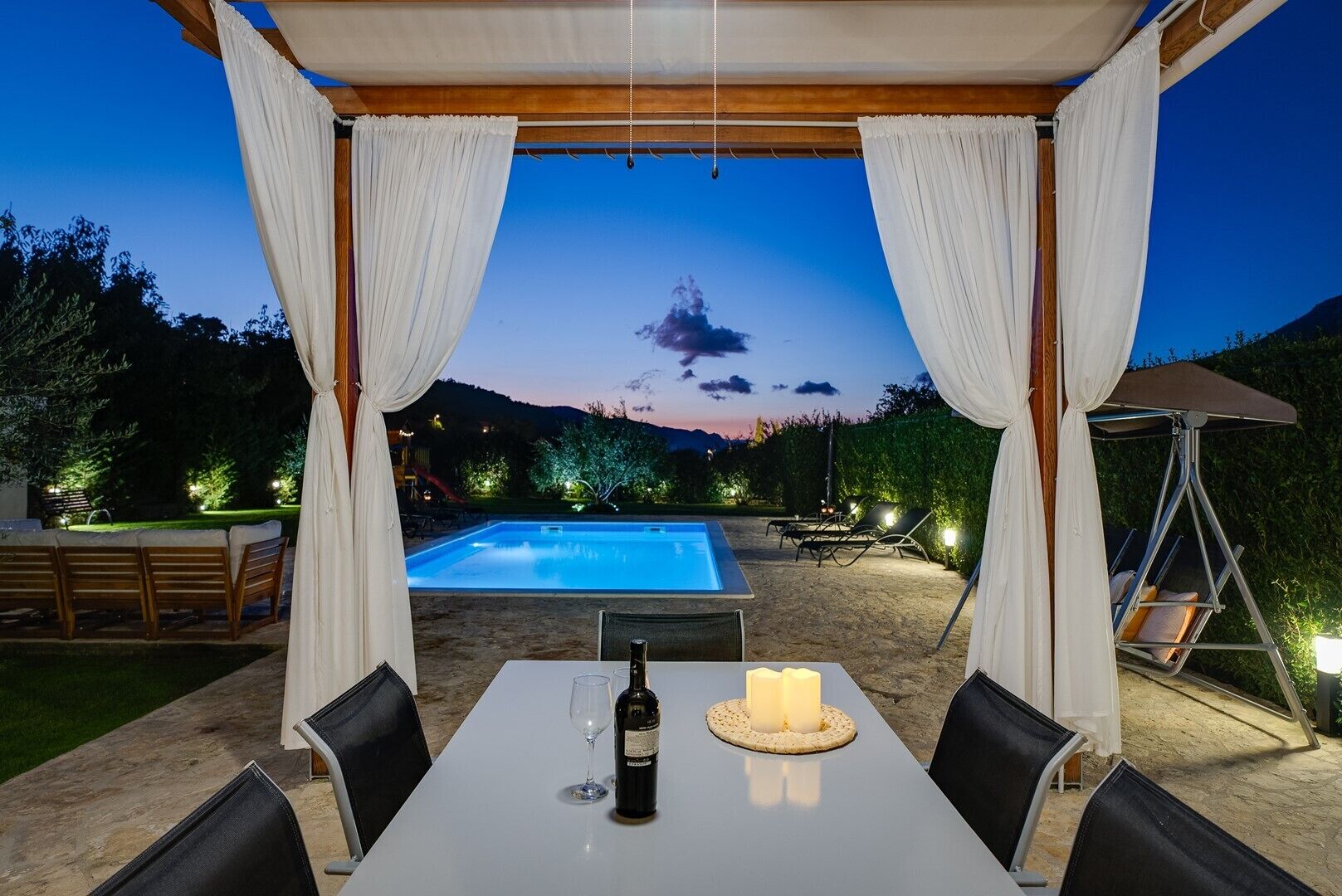 An outdoor dining area with a table, chairs, a bottle of wine, and candles. A pool is visible in the background.