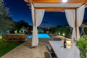 Une terrasse extérieure avec une piscine, un sol en pierre, des meubles en bois et un espace couvert agrémenté de rideaux blancs.