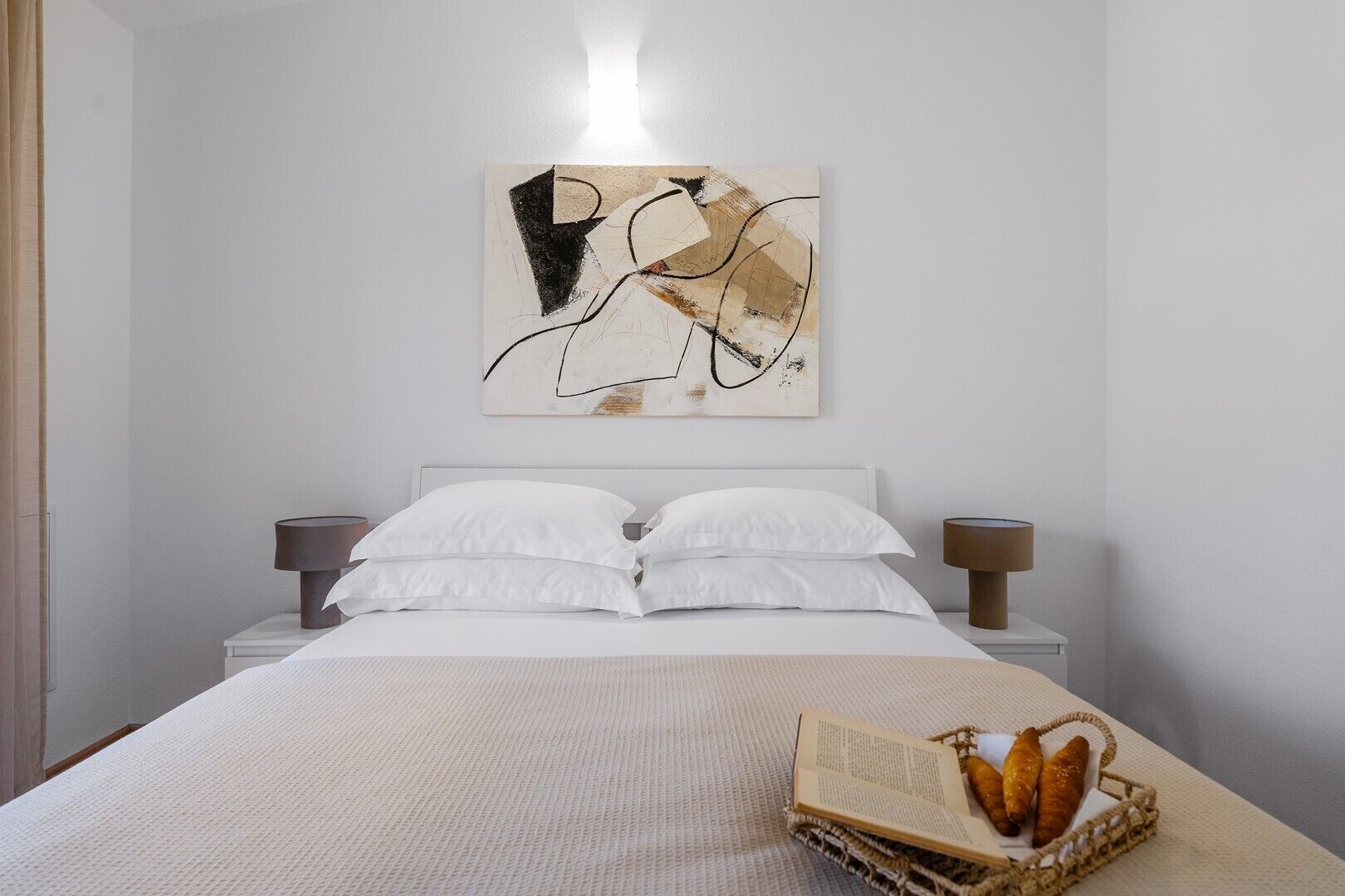 A neatly made bed with white linens and two bedside lamps, a wall art piece above the bed, and a basket with bread on the bed.