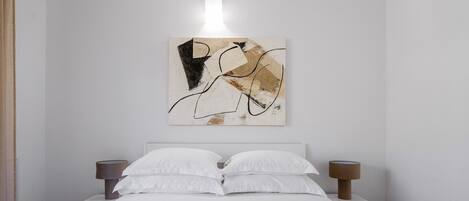A neatly made bed with white linens and two bedside lamps, a wall art piece above the bed, and a basket with bread on the bed.