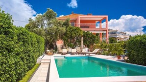 Un edificio de dos pisos con piscina, rodeado de vegetación y un cielo azul despejado.