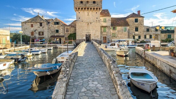 Un edificio de piedra con una torre, un camino de piedra que conduce al agua y varias embarcaciones amarradas a lo largo del canal.