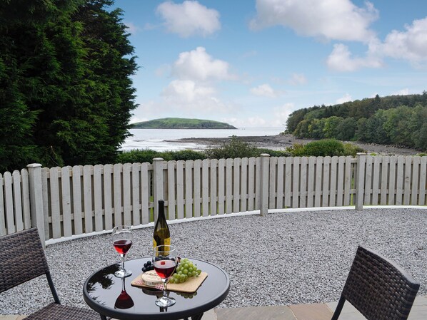 A table with a bottle, wine glasses, grapes, and cheese on a round table, set against a scenic backdrop of a beach and forest. - 1 bedroom accommodation in Balcary, near Auchencairn (Balcary, near Auchencairn)