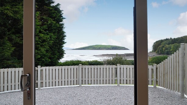 A view through a glass door of a gravel patio with a white picket fence, a body of water, and a green hill in the distance. - 1 bedroom accommodation in Balcary, near Auchencairn (Balcary, near Auchencairn)
