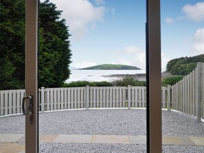 A view through a glass door of a gravel patio with a white picket fence, a body of water, and a green hill in the distance. - 1 bedroom accommodation in Balcary, near Auchencairn (Balcary, near Auchencairn)