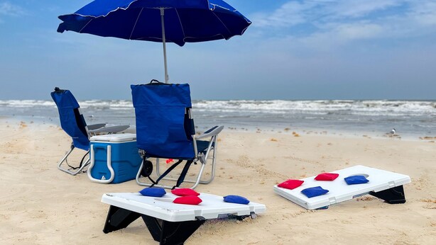 A beach setup with a blue umbrella, two beach chairs, a cooler, and a small white table with red and blue cushions. - Walk to Sandfest, Shared Pools, Boardwalk to the Beach (Port Aransas)
